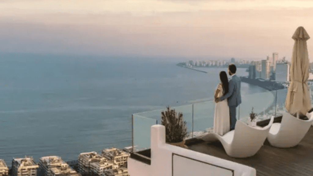 A couple standing on a luxury glass balcony at Prestige Ocean Towers looking out over the Arabian Sea at sunset.